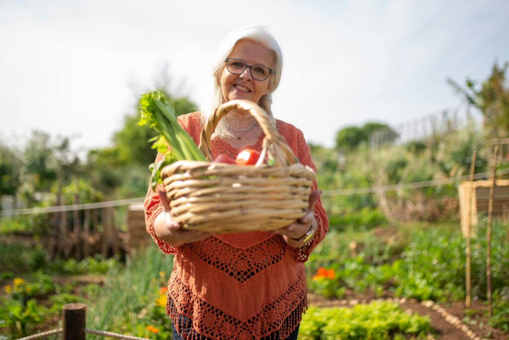 Elderly woman smiling with basket of fresh vegetables in a vibrant outdoor garden.